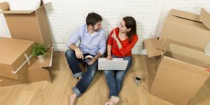 young happy American couple sitting on floor moving in a new house or apartment flat using computer laptop choosing online furniture and household in real estate and independent lifestyle concept