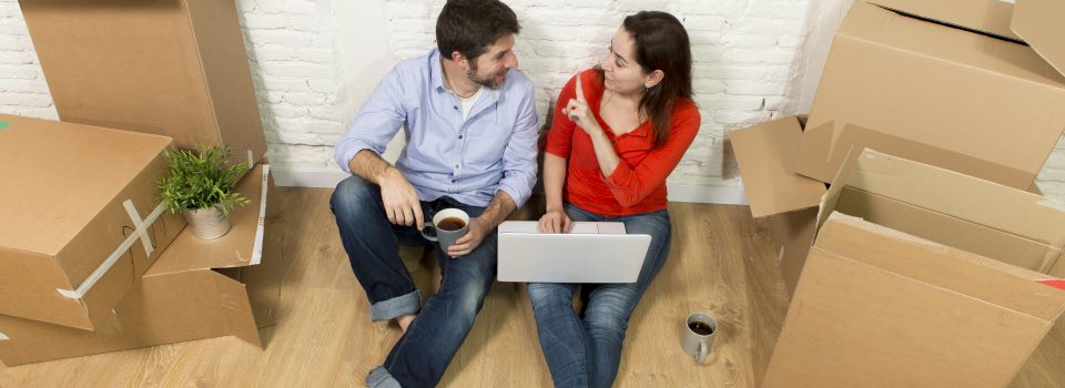 young happy American couple sitting on floor moving in a new house or apartment flat using computer laptop choosing online furniture and household in real estate and independent lifestyle concept